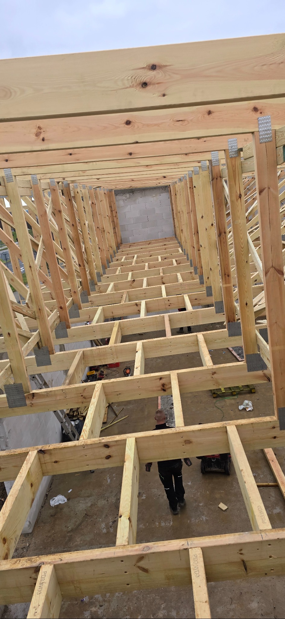 Wnętrze konstrukcji drewnianej dachu w trakcie budowy, widok z góry, z widocznymi elementami mocującymi i fragmentem osoby w dolnej części kadru.