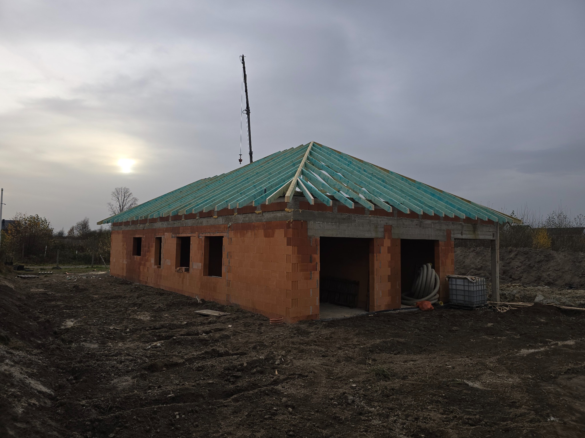 Budowa domu jednorodzinnego w Nowogrodźcu: ściany z czerwonej cegły, drewniana konstrukcja dachu z niebieską membraną, widoczny żuraw budowlany.