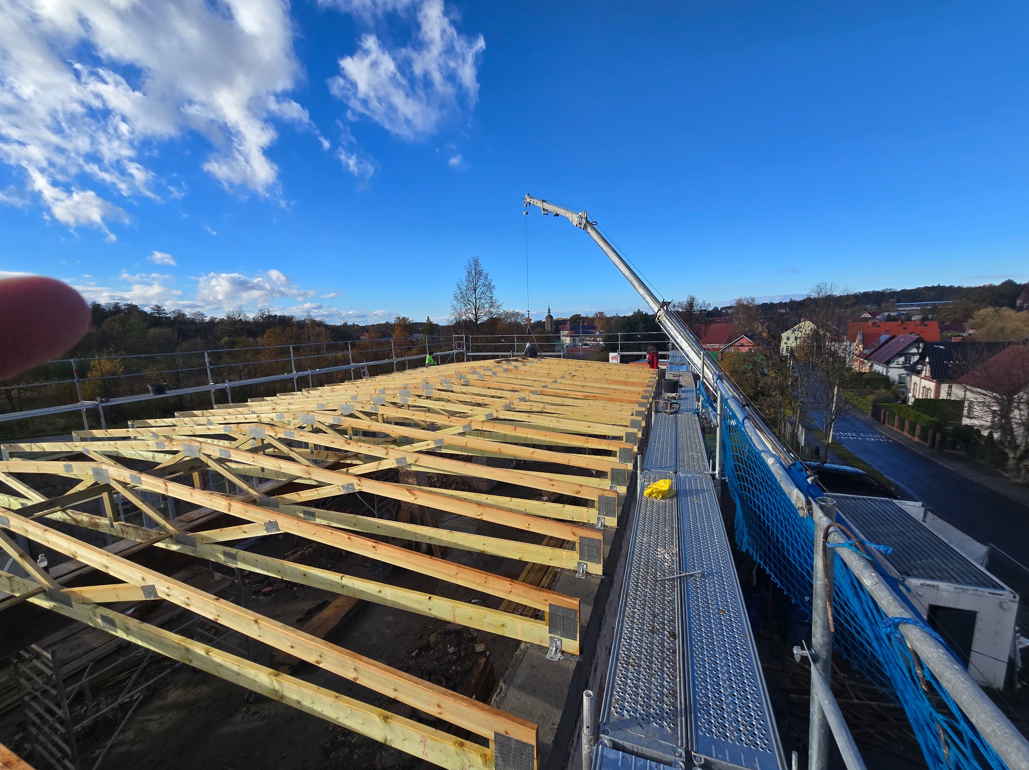 Wznoszenie drewnianej konstrukcji dachu z użyciem dźwigu na tle błękitnego nieba. Widoczne rusztowania i elementy zabezpieczeń.