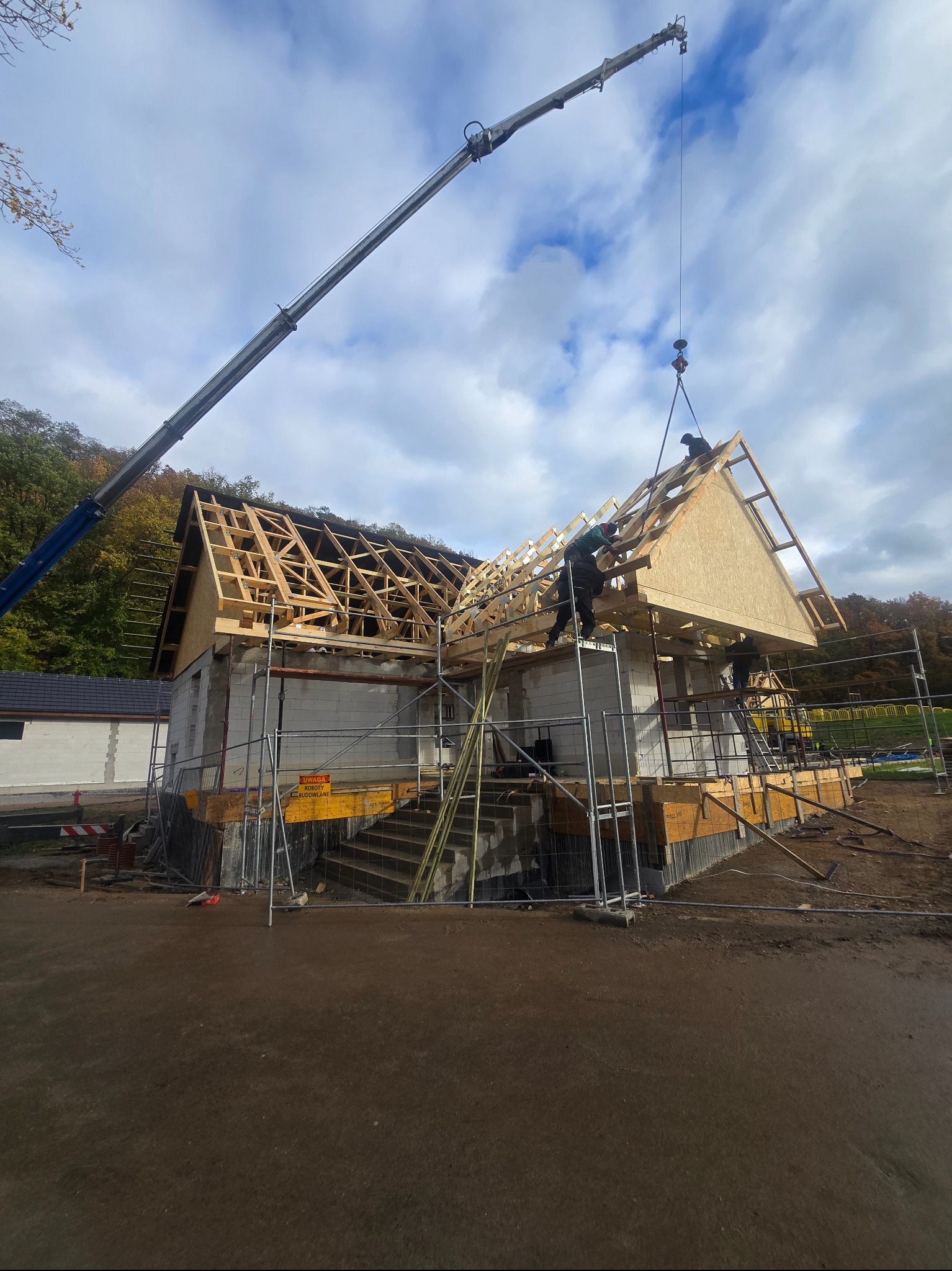 Montaż prefabrykowanej więźby dachowej na budynku z bloczków, z użyciem dźwigu. Widoczni pracownicy na rusztowaniach. Tło: drzewa i pochmurne niebo.