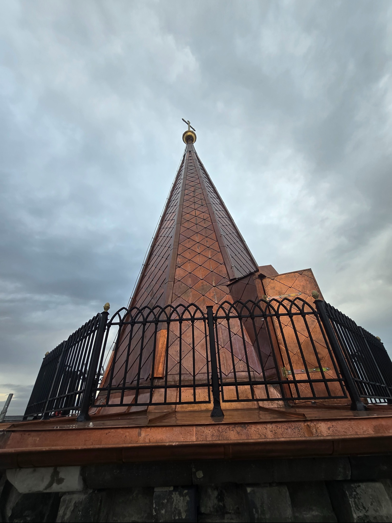 Miedziana wieża kościoła z krzyżem na szczycie, widoczna z dołu, otoczona czarnym, ozdobnym ogrodzeniem na dachu. Pochmurne niebo w tle.
