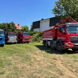 Ciężar&oacute;wki budowlane r&oacute;żnych marek (MAN, Scania) zaparkowane na trawiastym terenie z koparką w tle.