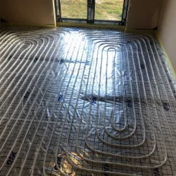 Białe rurki systemu grzewczego podłogowego ułożone w pętle na srebrnej folii z nadrukiem siatki, przygotowane do zalania wylewką, widoczne okno z ramą VEKA i żółta taśma dylatacyjna przy ścianach.