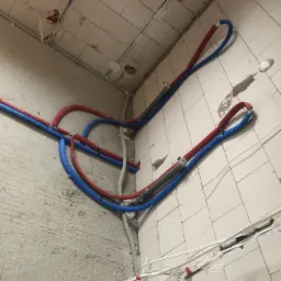 Instalacja rur wodnych (niebieskie i czerwone) oraz przewod&oacute;w elektrycznych w surowym wnętrzu z częściowo wykończonymi ścianami z białych płytek.