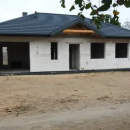 Niedokończony dom murowany z białych bloczków, z ciemnoszarą dachówką, widoczne otwory okienne i drzwiowe, teren wokół budynku piaszczysty.