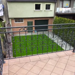 Kuta, czarna balustrada balkonowa z ozdobnymi elementami na tle zadbanego trawnika i budynku z garażem. Widok z góry na płytki balkonowe.