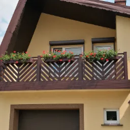 Balkon z brązową, drewnianą konstrukcją z motywem jodełki, ozdobiony skrzynkami z czerwonymi kwiatami na tle beżowej elewacji domu z brązowym dachem.