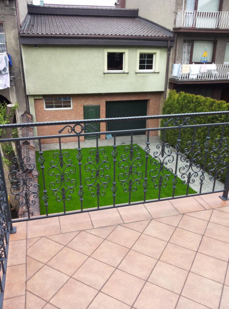 Kuta, czarna balustrada balkonowa z ozdobnymi elementami na tle zadbanego trawnika i budynku z garażem. Widok z góry na płytki balkonowe.