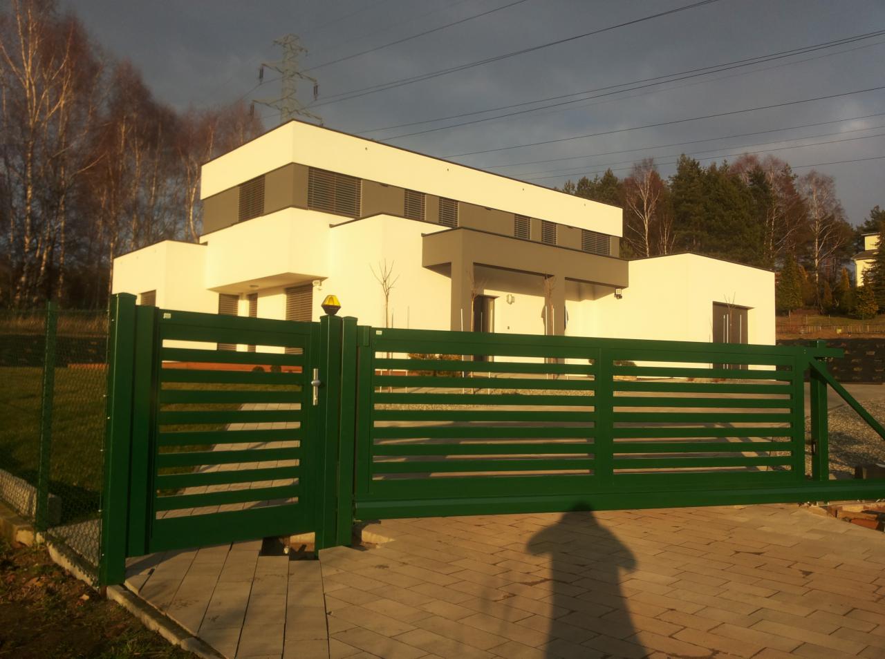 Nowoczesna, pozioma, zielona brama przesuwna z automatyką, z widocznym cieniem osoby na kostce brukowej, na tle białego domu z szarymi elementami.