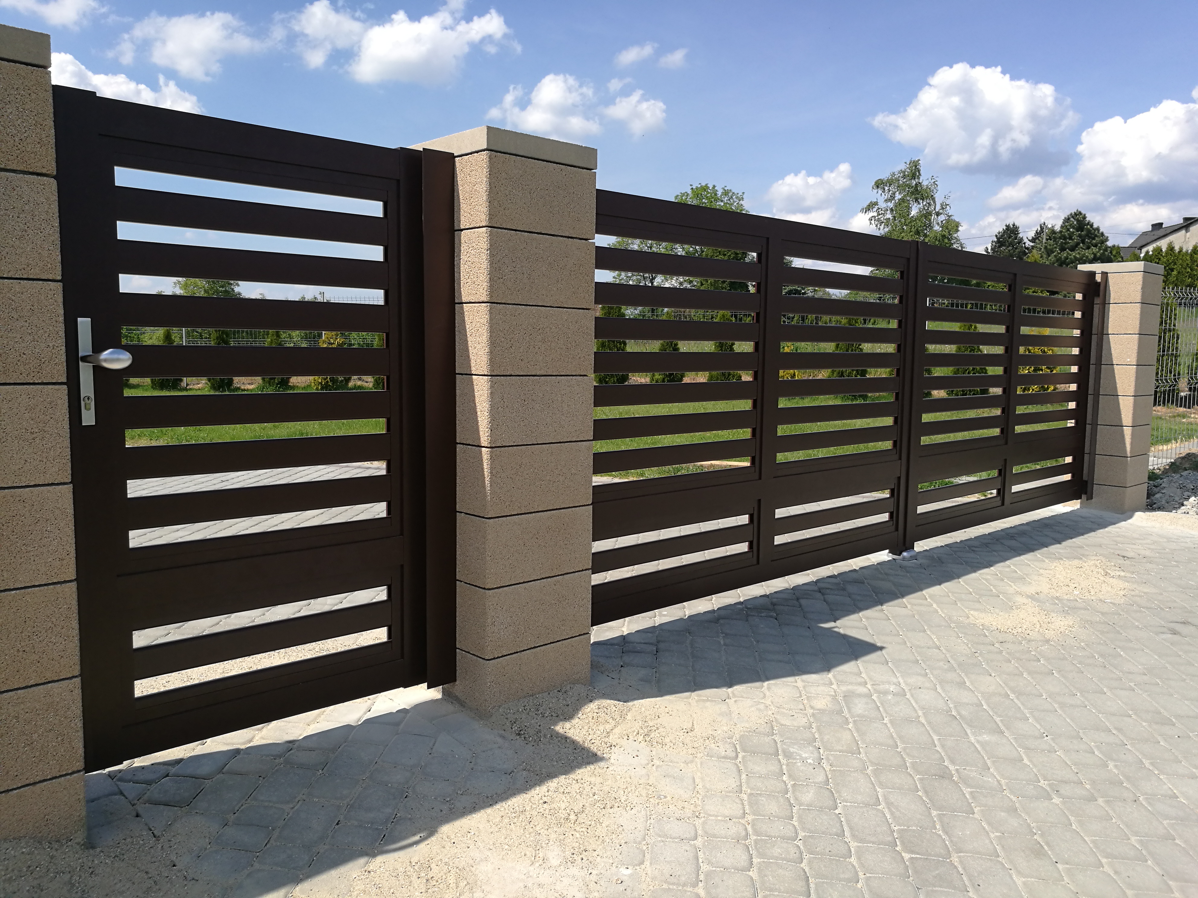 Nowoczesna brama wjazdowa z poziomymi przęsłami w kolorze ciemnego brązu, zamontowana na słupkach z szarego betonu architektonicznego w Rybniku, widok z zewnątrz na kostkę brukową i trawnik.