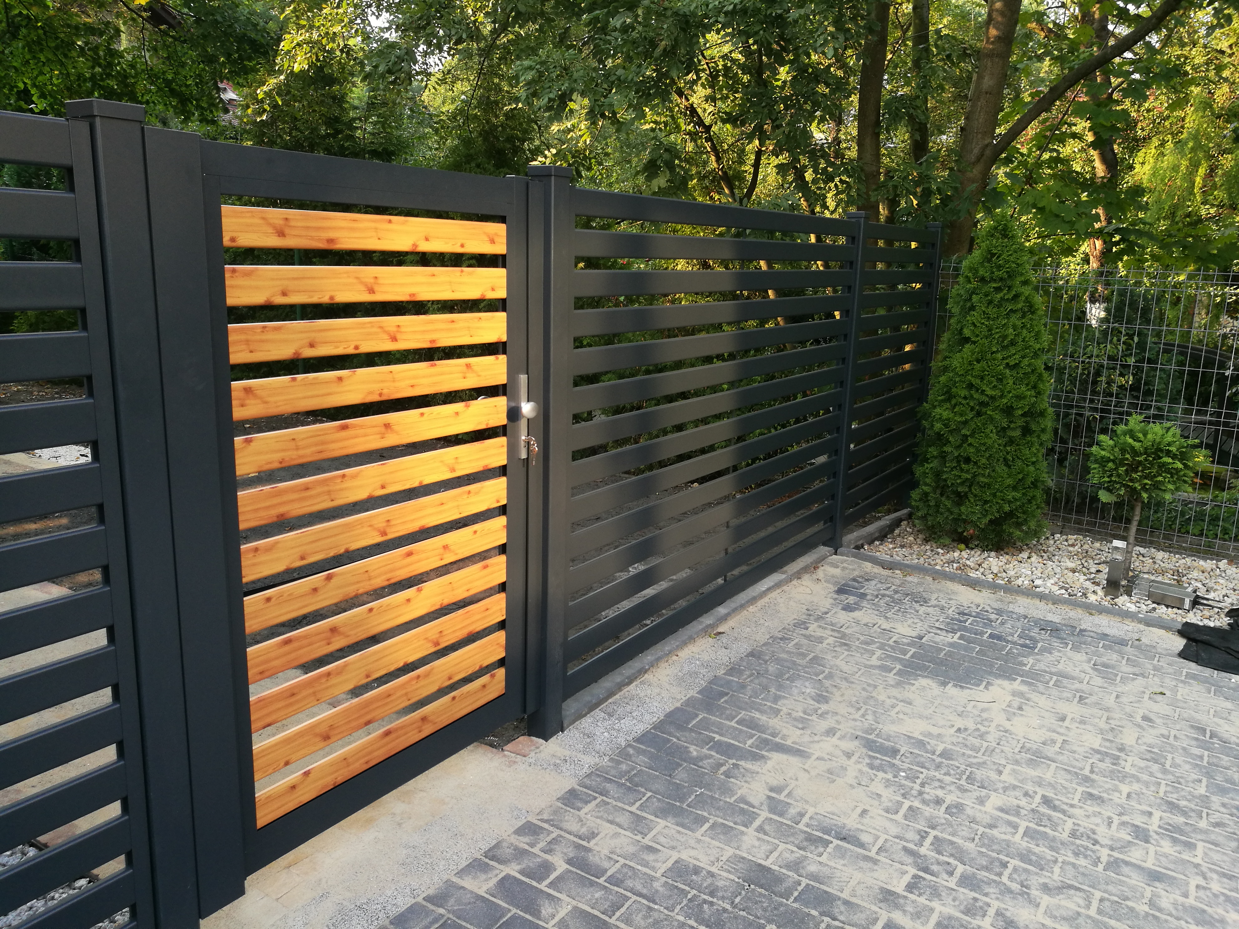 Nowoczesne ogrodzenie posesji z przesuwną bramą, wykonane z szarych, metalowych profili i drewnianych paneli w kolorze jasnego drewna, widoczne z zewnątrz na tle zieleni.