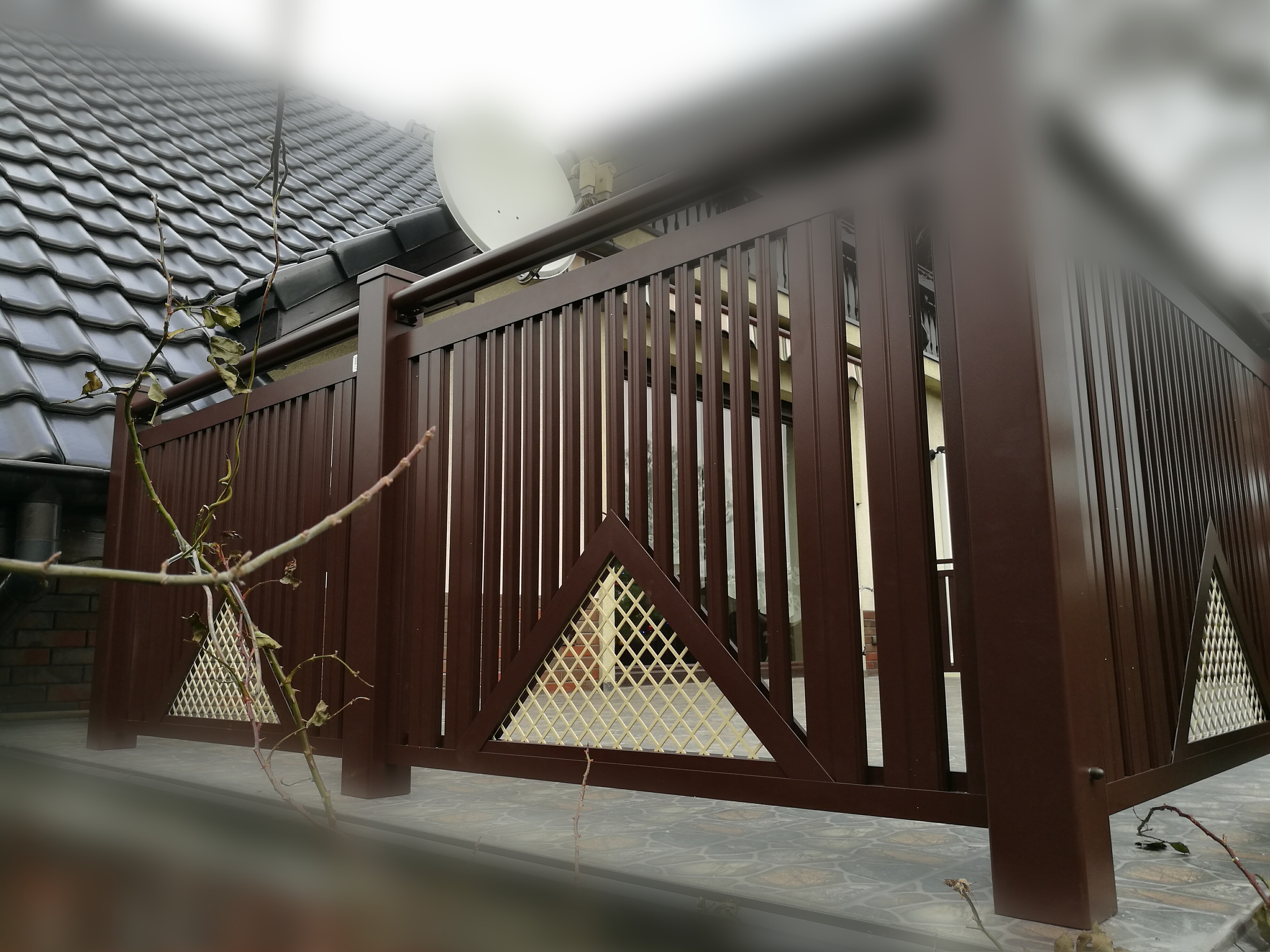 Ciemnobrązowe ogrodzenie z pionowych paneli, z trójkątnymi wstawkami z siatki, widoczne z bliska, z tłem budynku i anteny satelitarnej.