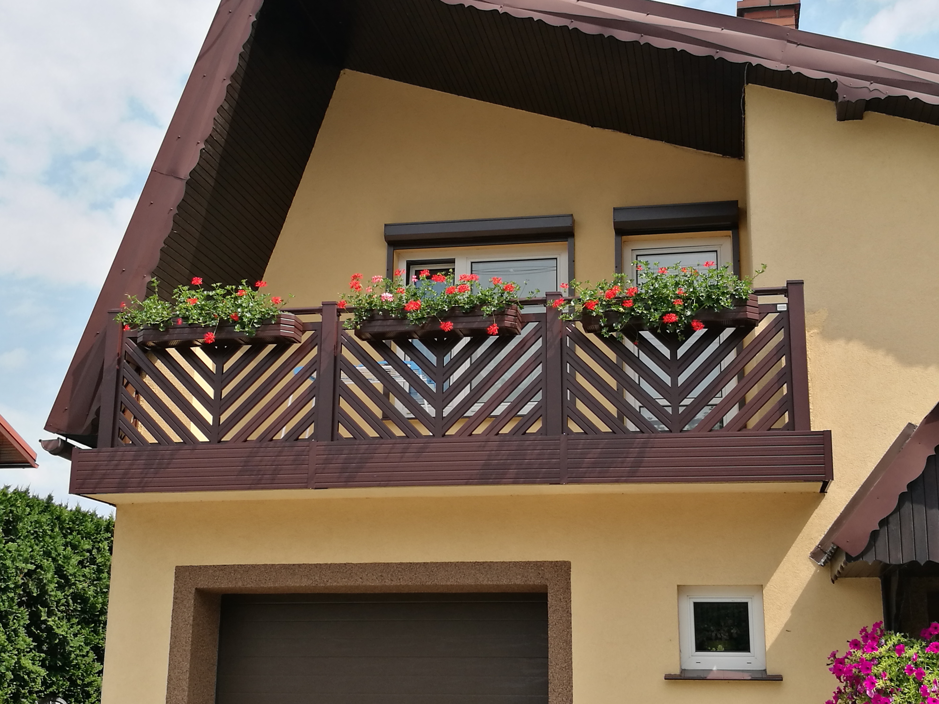 Balkon z brązową, drewnianą konstrukcją z motywem jodełki, ozdobiony skrzynkami z czerwonymi kwiatami na tle beżowej elewacji domu z brązowym dachem.