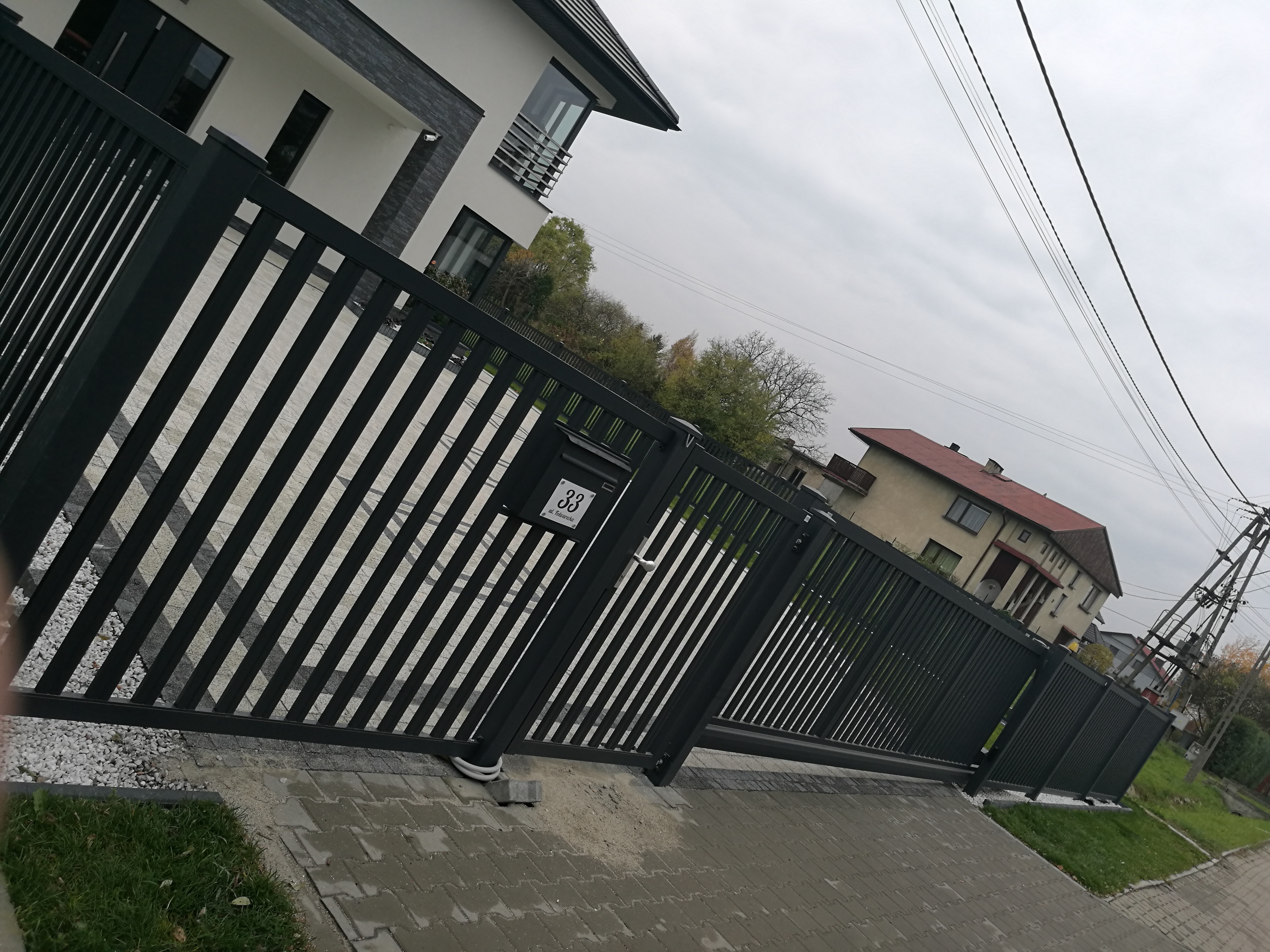 Nowoczesne ogrodzenie posesji z czarnych, pionowych profili, widoczna skrzynka na listy z numerem 33, w tle dom z białymi ścianami i szarymi elementami dekoracyjnymi.