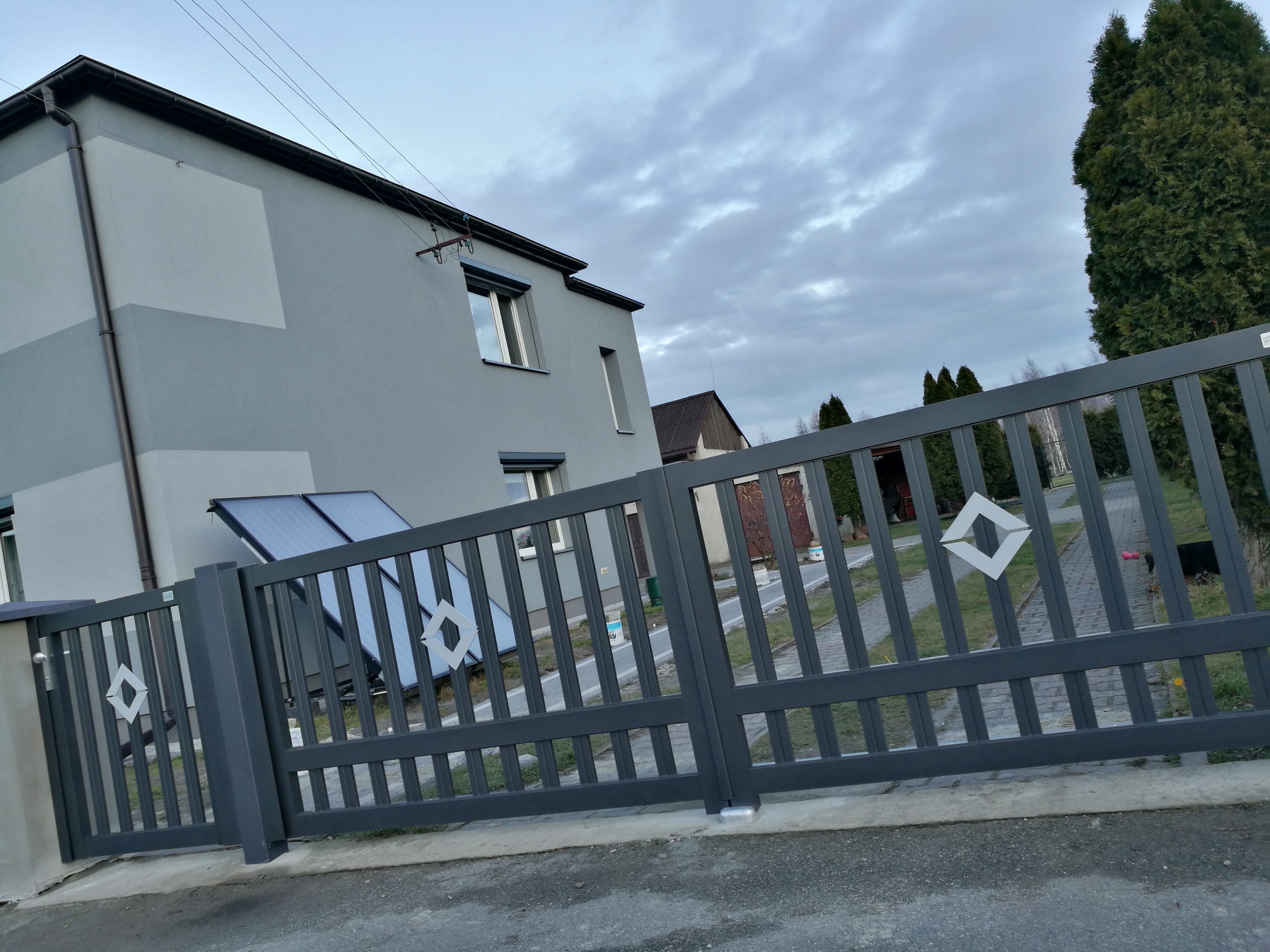 Nowoczesna, grafitowa brama posesyjna z elementami dekoracyjnymi w kształcie rombów, na tle domu z panelami solarnymi i brukowanym podjazdem.