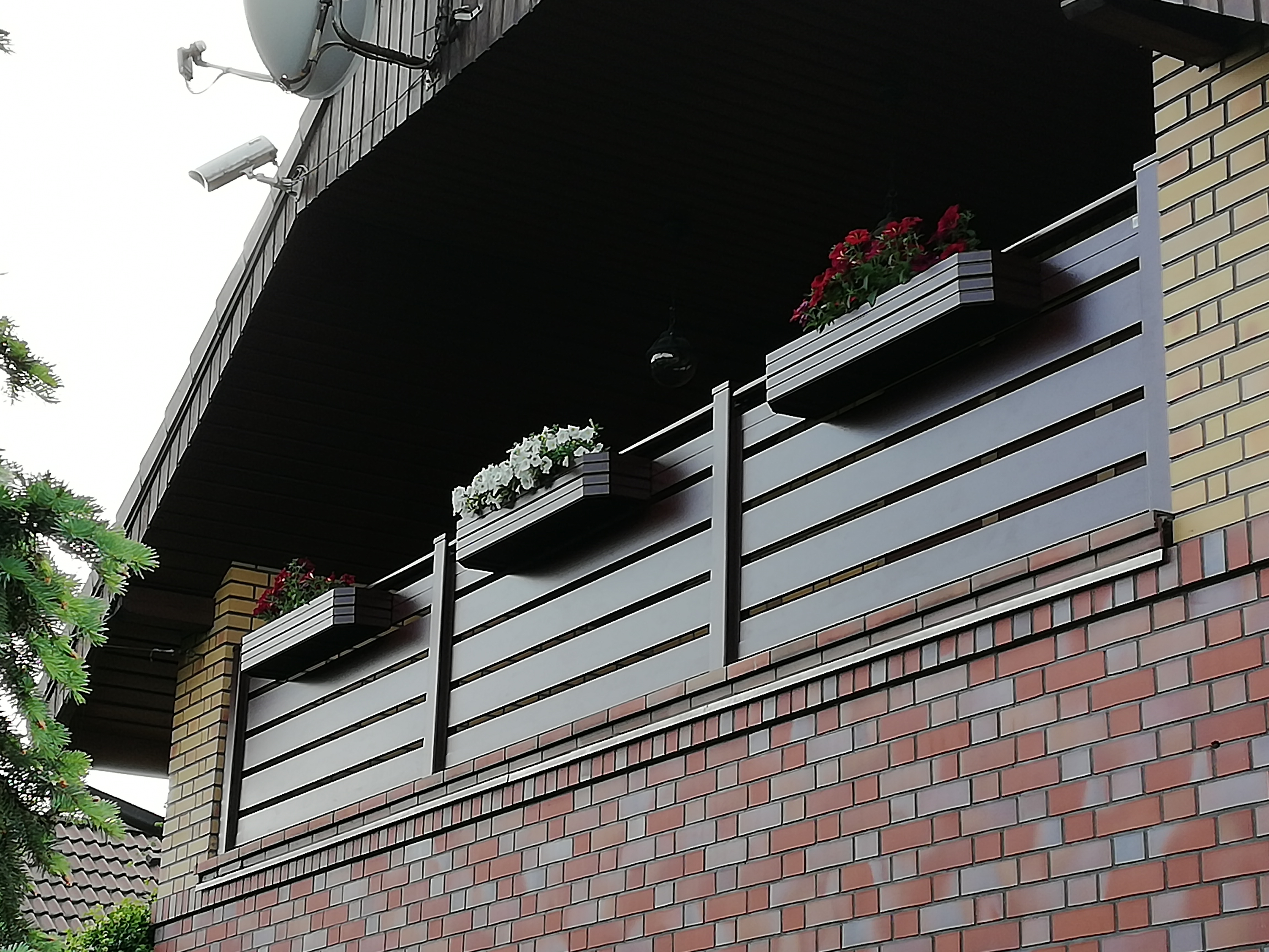 Balkon z drewnopodobną osłoną, zdobiony trzema skrzynkami z kwiatami: czerwonymi, białymi i ponownie czerwonymi. Widoczna antena satelitarna i kamera monitoringu na dachu.