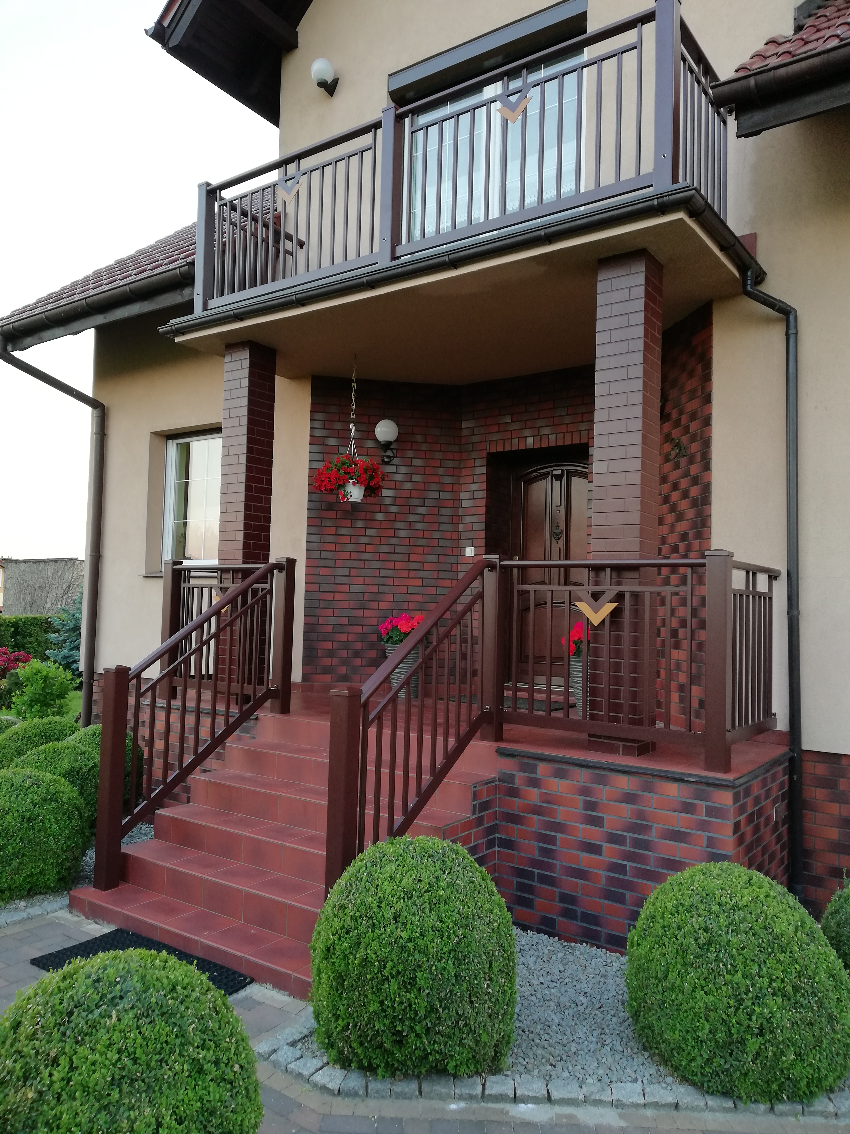 Brązowe balustrady metalowe przy schodach wejściowych do domu z ceglaną elewacją i balkonem, ozdobione kwiatami w donicach.