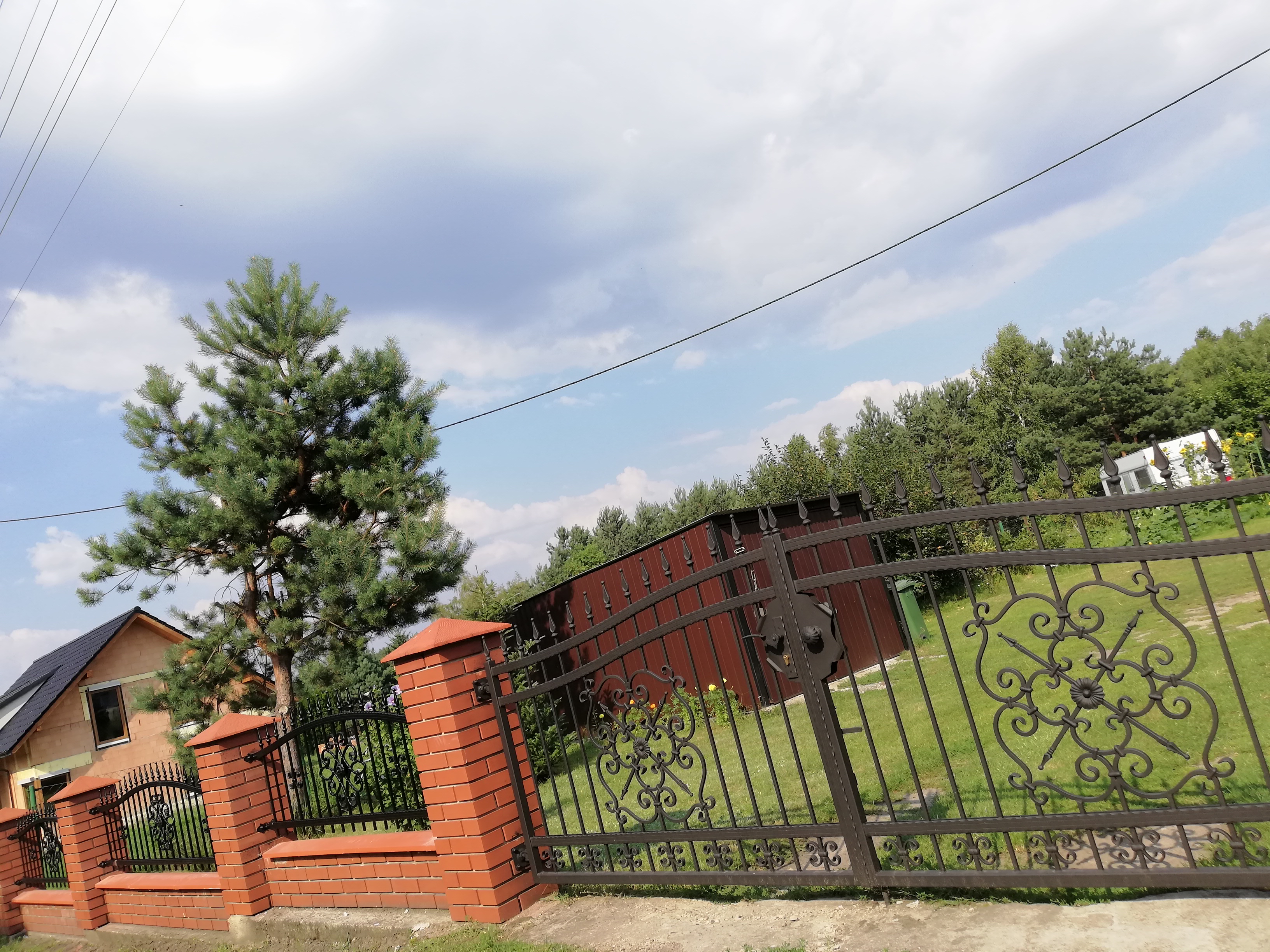 Kute ogrodzenie z bramą, słupki z czerwonej cegły, w tle dom w trakcie budowy i wysokie drzewo iglaste.