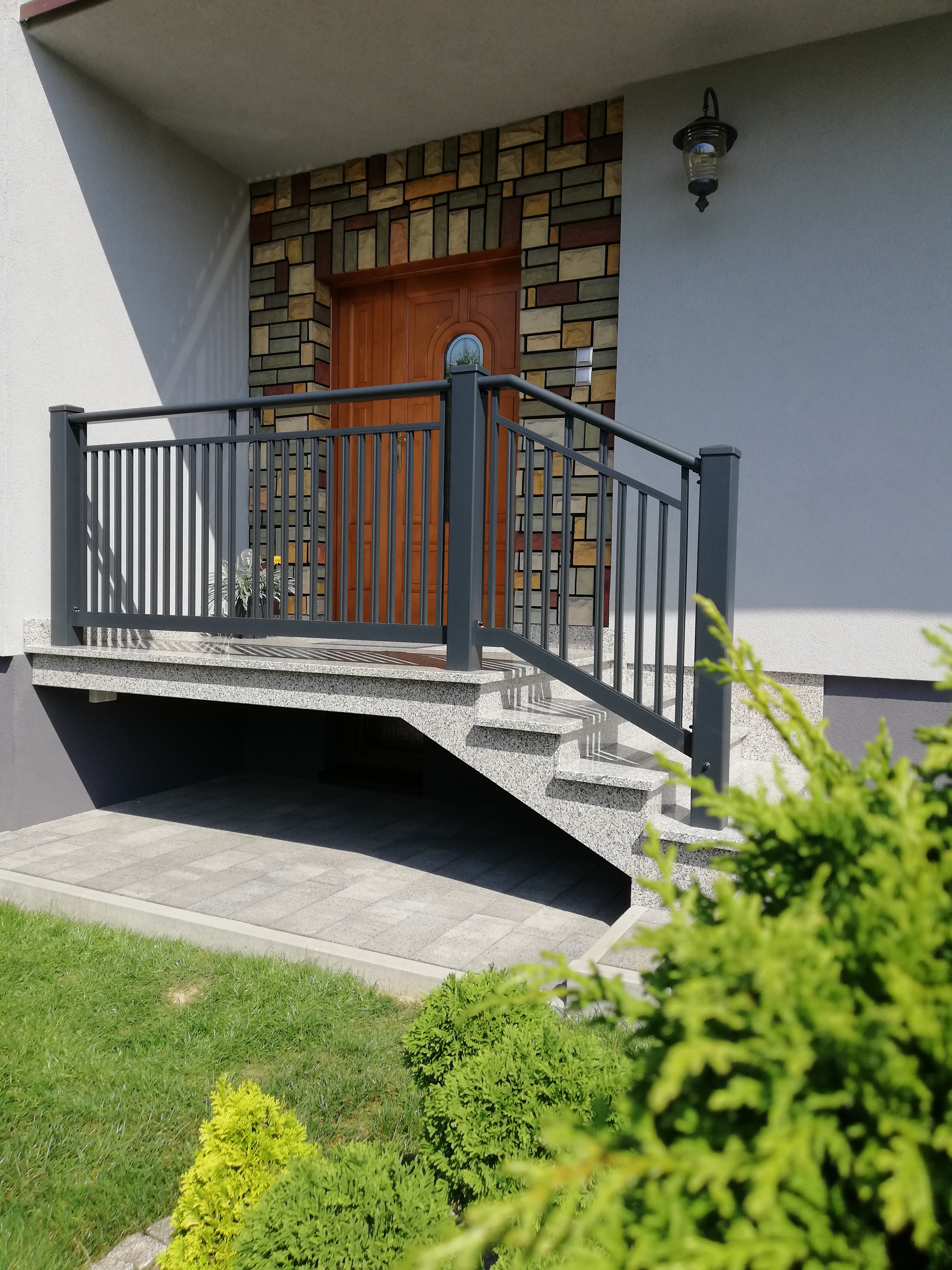 Zewnętrzne schody wejściowe z szarej, metalowej balustradą, granitowe stopnie, drzwi wejściowe z okładziną z kamienia dekoracyjnego, lampa ścienna, widok z poziomu trawnika.