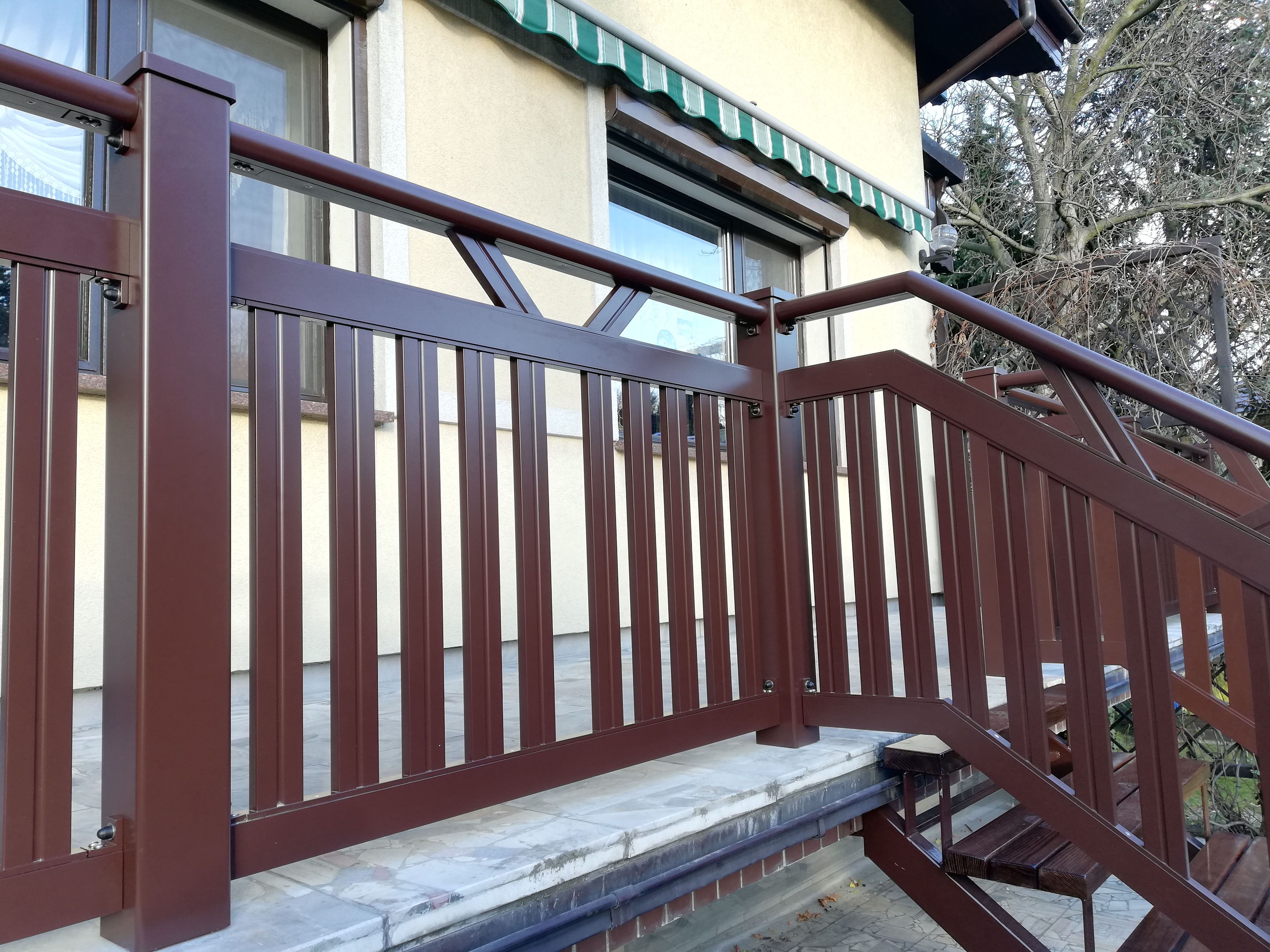 Zewnętrzne schody z brązową metalową konstrukcją i poręczą, widoczne okna i markiza w tle.