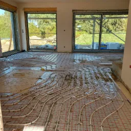 Białe rurki ogrzewania podłogowego rozłożone na czerwonej siatce w surowym wnętrzu domu w trakcie budowy, widok przez niedokończone przejście między pomieszczeniami.