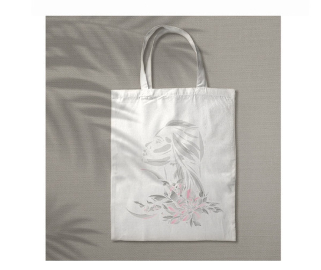 Biała torba bawełniana z nadrukiem graficznym przedstawiającym profil kobiety z kwiatami w odcieniach szarości i różu, leżąca na szarym tle z cieniem liści.