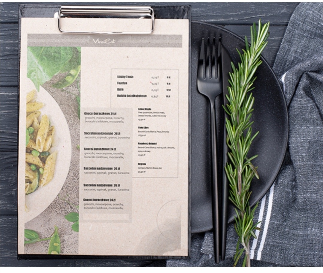 Menu restauracji z ilustracją dania z makaronu, czarny talerz z widelcem i gałązką rozmarynu na tle drewnianego stołu i szarej ściereczki.