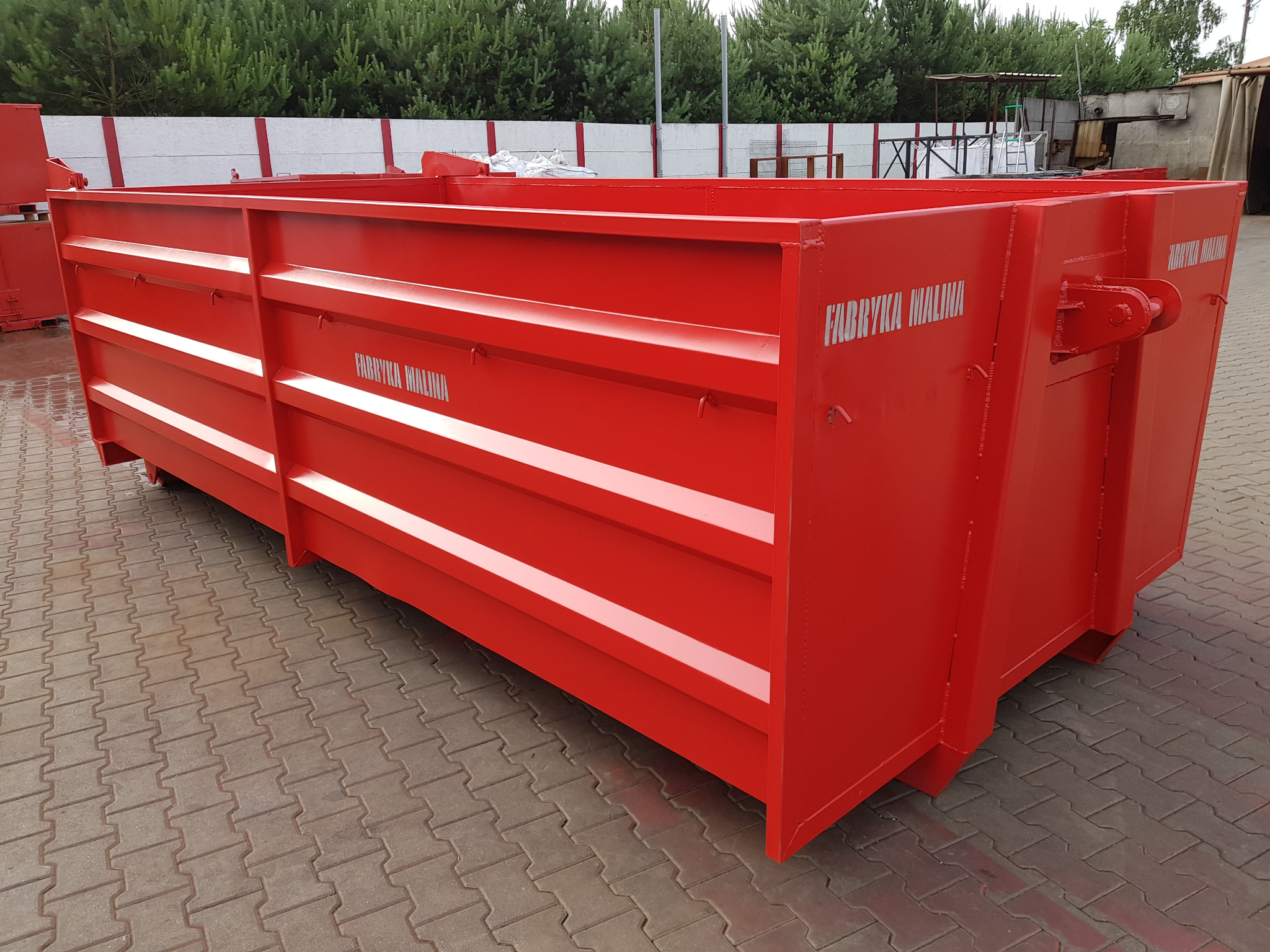 Czerwony, metalowy kontener z napisem 'Fabryka Malina' na kostce brukowej, widok z boku.
