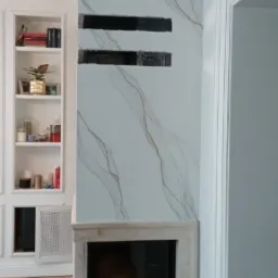 Nowoczesny kominek obudowany płytami imitującymi marmur z czarnymi, poziomymi akcentami dekoracyjnymi, wkomponowany w ścianę z zabudową meblową w tle.