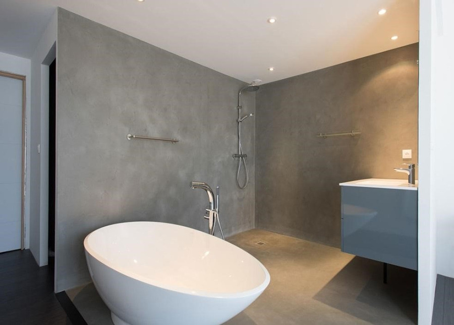 Minimalistyczna łazienka z szarą, gładką powierzchnią ścian i podłogi, wolnostojąca biała wanna o owalnym kształcie, chromowana armatura, szary szafka z umywalką oraz punktowe oświetlenie sufitowe.