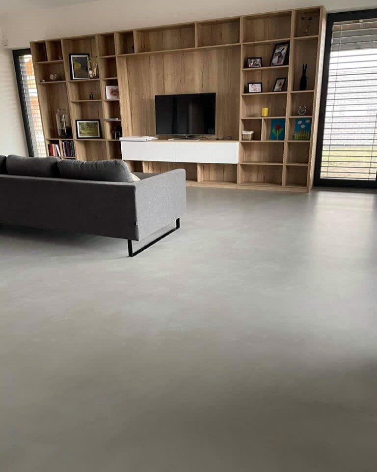 Szara, jednolita podłoga w salonie z dużym regałem na książki i szarą sofą. Widoczne delikatne refleksy światła na gładkiej powierzchni podłogi.