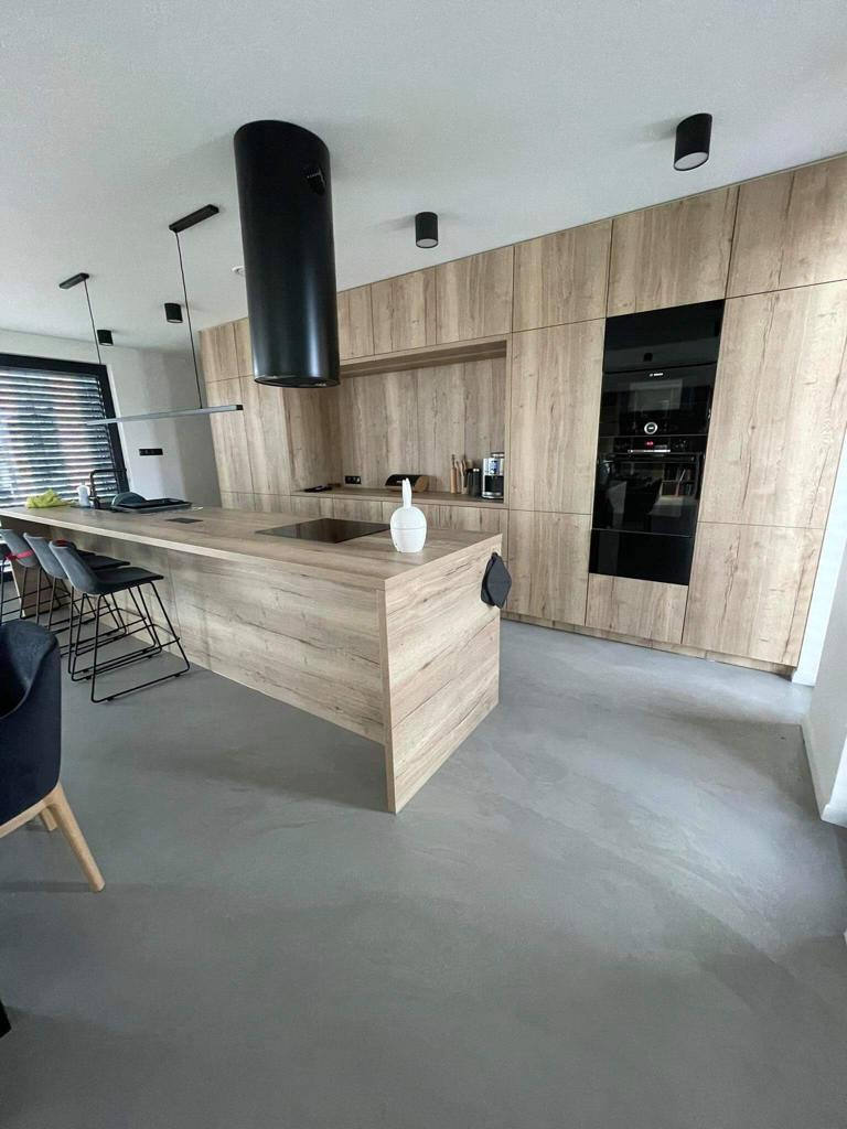 Nowoczesna kuchnia z wyspą i posadzką żywiczną w odcieniach szarości, drewniane fronty szafek, czarny okap cylindryczny i wbudowany piekarnik, minimalistyczne oświetlenie.