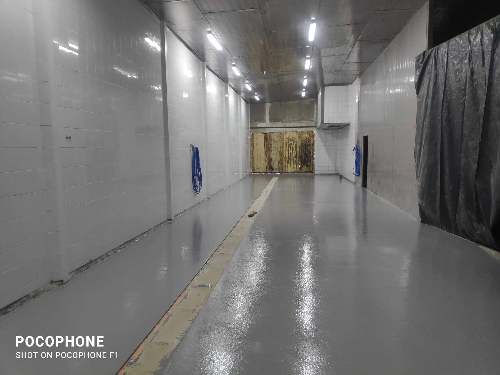 Wnętrze hali z nową, jednolitą, szarą posadzką, białe kafelki na ścianach, widoczne rury pod sufitem i zasłonięty fragment przestrzeni po prawej stronie.