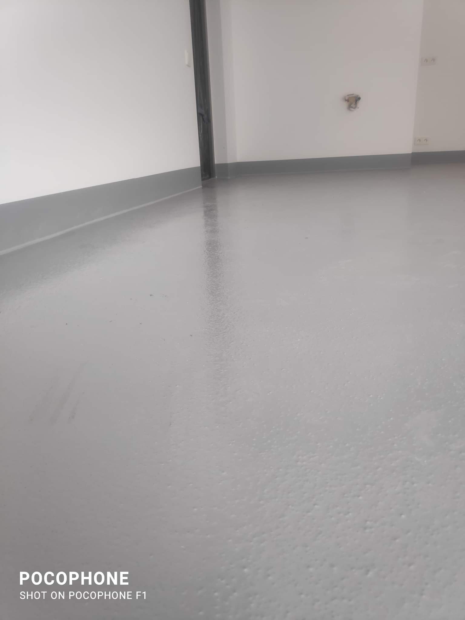 Gładka, szara podłoga w pomieszczeniu z białymi ścianami i szarą listwą przypodłogową, widoczne ślady po niedawnej aplikacji, fragment ściany z widocznym gniazdkiem elektrycznym.