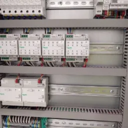 Wewnętrzny widok szarej szafy sterowniczej z zamontowanymi modułami elektrycznymi Schneider Electric, widoczne przewody i listwy zaciskowe.
