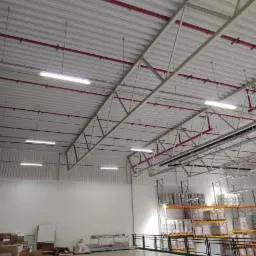 Wnętrze hali magazynowej z widocznymi rzędami lamp LED zamontowanymi pod sufitem i systemem rur przeciwpożarowych w kolorze czerwonym.