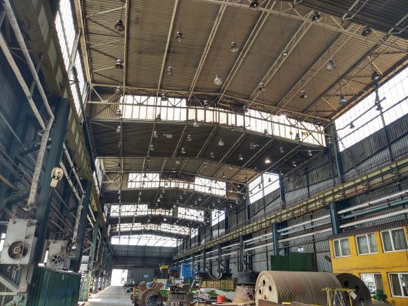 Wnętrze hali przemysłowej z widocznymi elementami konstrukcyjnymi, oświetleniem sufitowym i wyposażeniem technicznym. Widoczne liczne lampy przemysłowe zawieszone pod sufitem. Elementy...