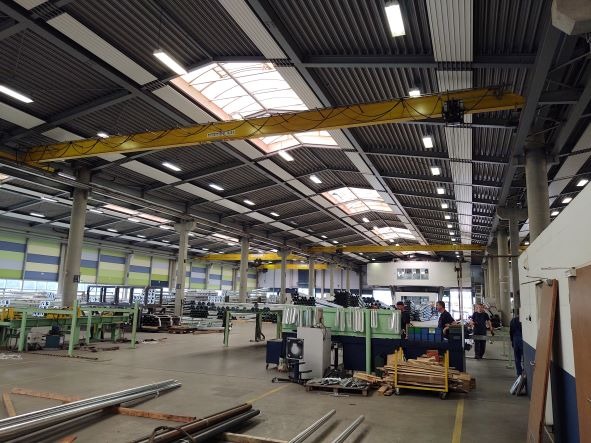 Wnętrze hali produkcyjnej z suwnicą pomostową w kolorze żółtym pod sufitem, oświetlonej lampami i światłem dziennym z okien dachowych.