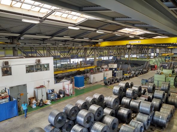 Widok z góry na halę produkcyjną z suwnicą, rolkami blachy i pracownikiem w niebieskiej koszulce.