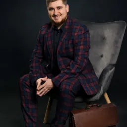 Uśmiechnięty mężczyzna z brodą w kraciastej marynarce i spodniach, siedzący na szarym fotelu obok brązowej skórzanej aktówki, na ciemnym tle.