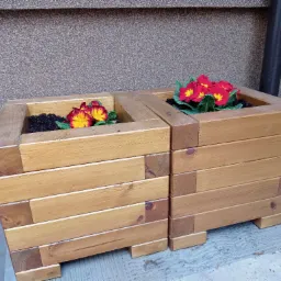 Dwie drewniane donice skrzyniowe z czerwono-żółtymi bratkami, wykonane z poziomych desek, stojące na betonowym podłożu przy szarej ścianie.