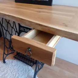 Drewniany blat biurka z wysuwaną szufladą i nogami od starej maszyny do szycia Singer, ustawiony na puszystym dywaniku.