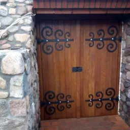 Drewniane drzwi wejściowe z ozdobnymi, kutymi zawiasami, wkomponowane w kamienną ścianę z ceglanym nadprożem.