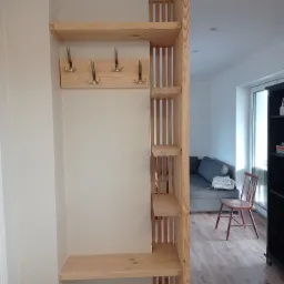 Drewniana zabudowa przedpokoju z półkami i wieszakami, jasne drewno, minimalistyczny design, widok na salon z szarą sofą.