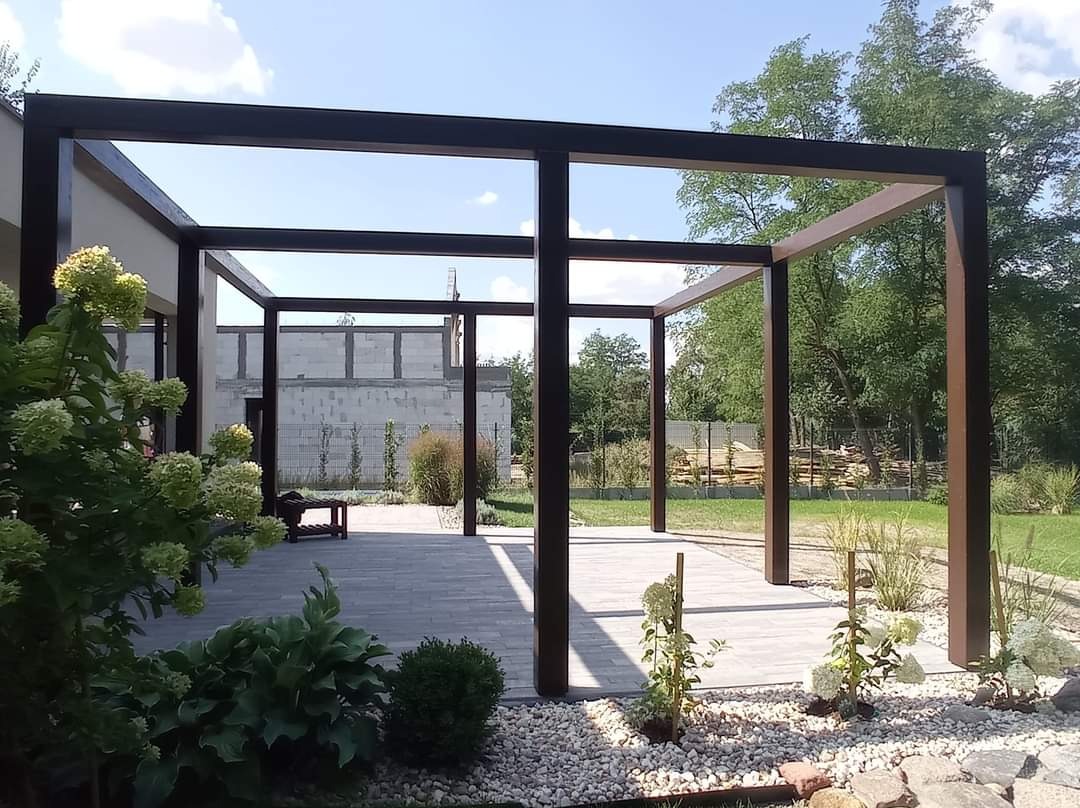 Nowoczesna, minimalistyczna pergola z ciemnego drewna na tarasie z szarej kostki, otoczona zielenią i białymi kamieniami, w tle budynek w trakcie budowy.