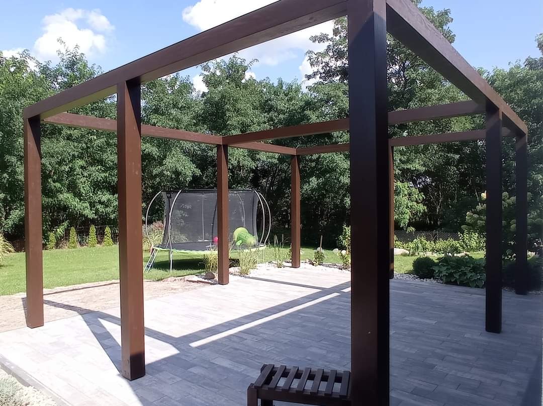 Brązowa, drewniana konstrukcja tarasu z widocznym trampoliną w tle, ustawiona na szarej kostce brukowej w słoneczny dzień.