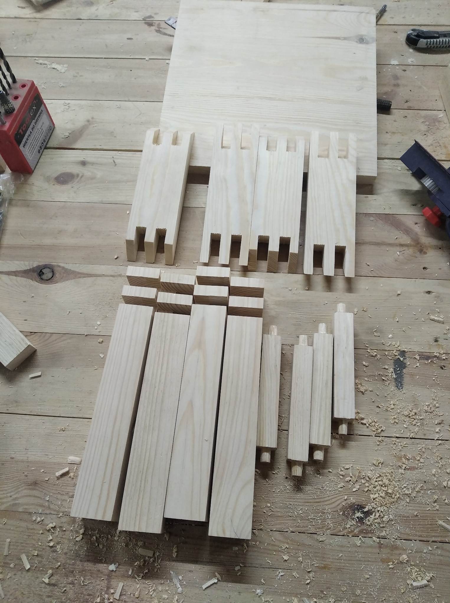 Przygotowane elementy z jasnego drewna do montażu mebli, widoczne czopy i wpusty, wiertła oraz ślady obróbki na drewnianym blacie roboczym.