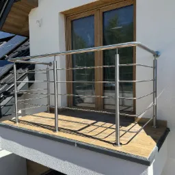 Balkon z balustradą ze stali nierdzewnej, drewnopodobne płytki na podłodze, okno z drewnianą ramą, panele słoneczne na dachu. Nowoczesne wykończenie.