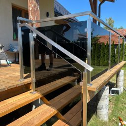 Modimet - Szklana balustrada z metalowymi słupkami na drewnianym tarasie i schodach. Widoczne odbicie osoby. Tło: dom z ogrodem, słoneczny dzień.