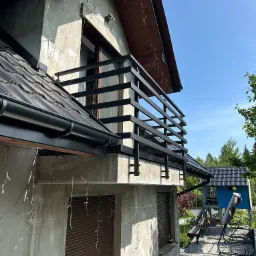 Czarna, pozioma balustrada balkonowa z metalu na tle jasnej elewacji domu i błękitnego nieba. Widok z dołu, oświetlenie słoneczne.